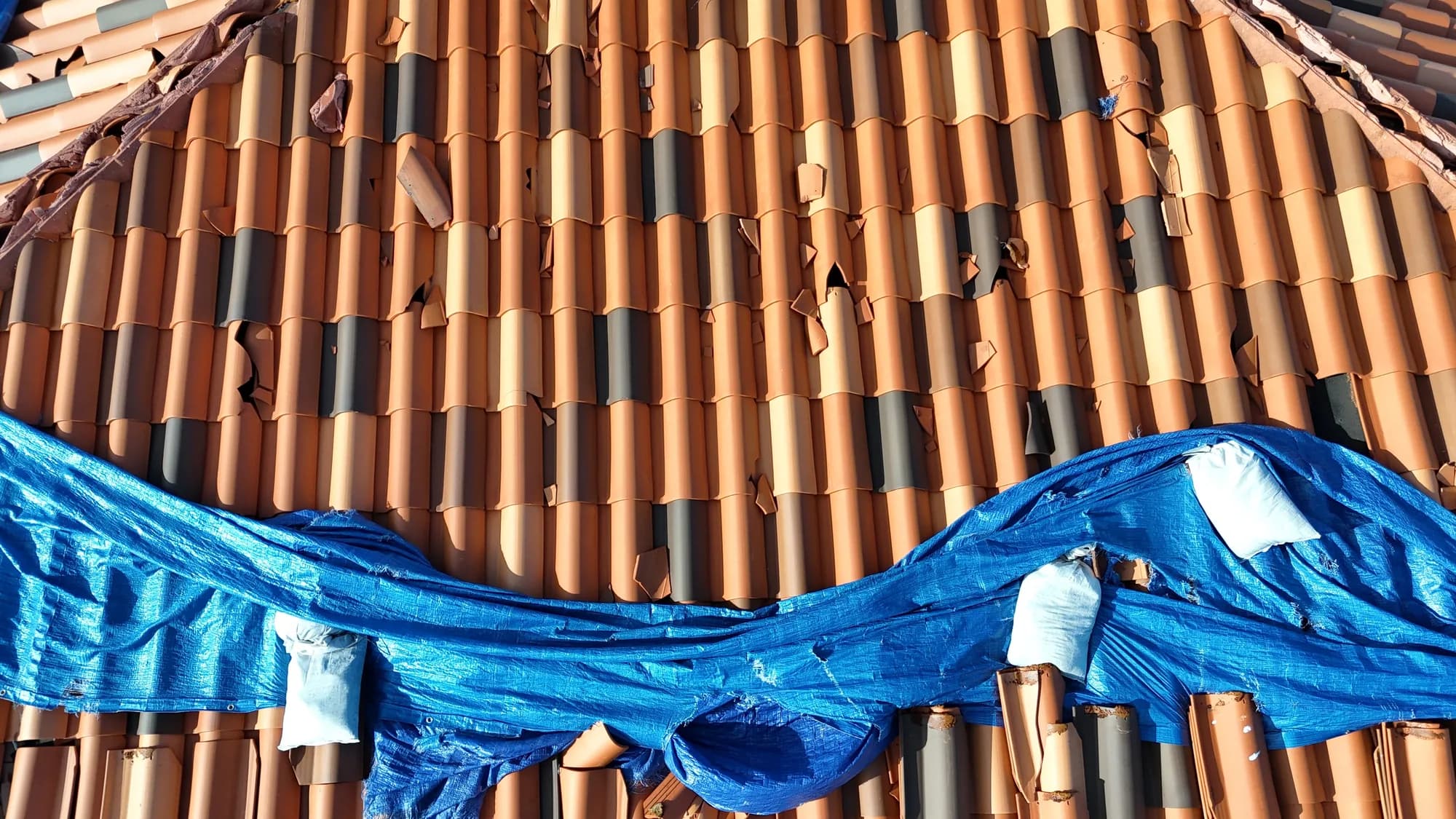 Drone view of storm-damaged clay tile and torn tarp material across a roof slope