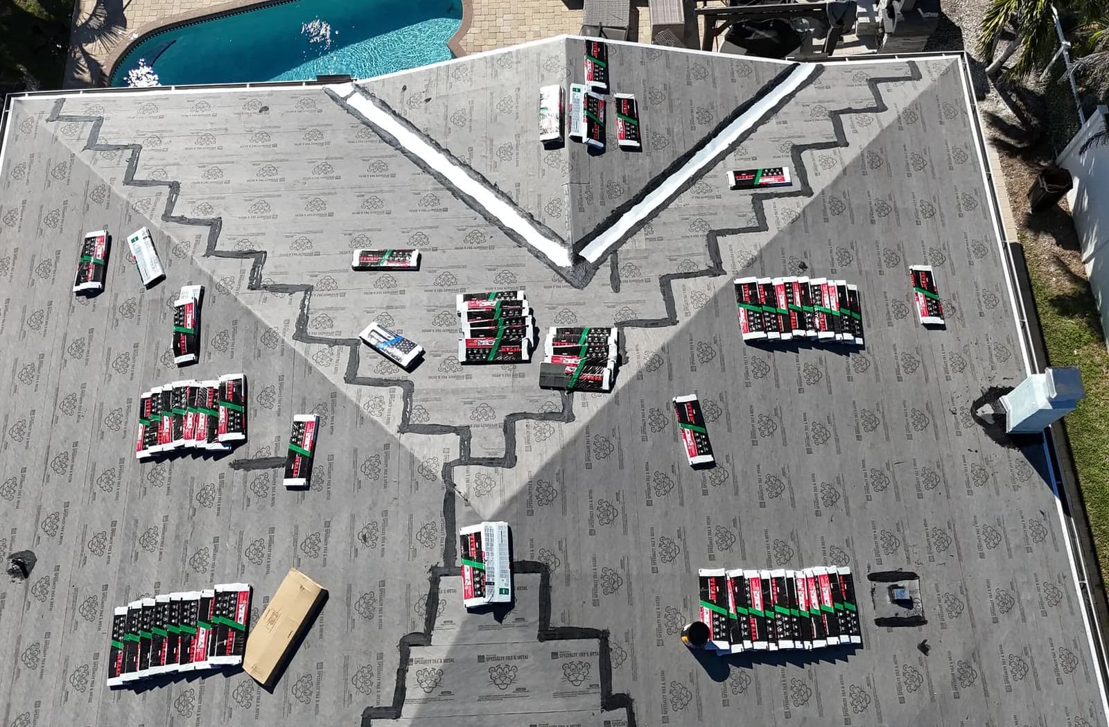 Aerial view of a hip roof fully dried in with grey underlayment, seams taped and sealed, shingle bundles staged across the slopes, and a pool beside the house.