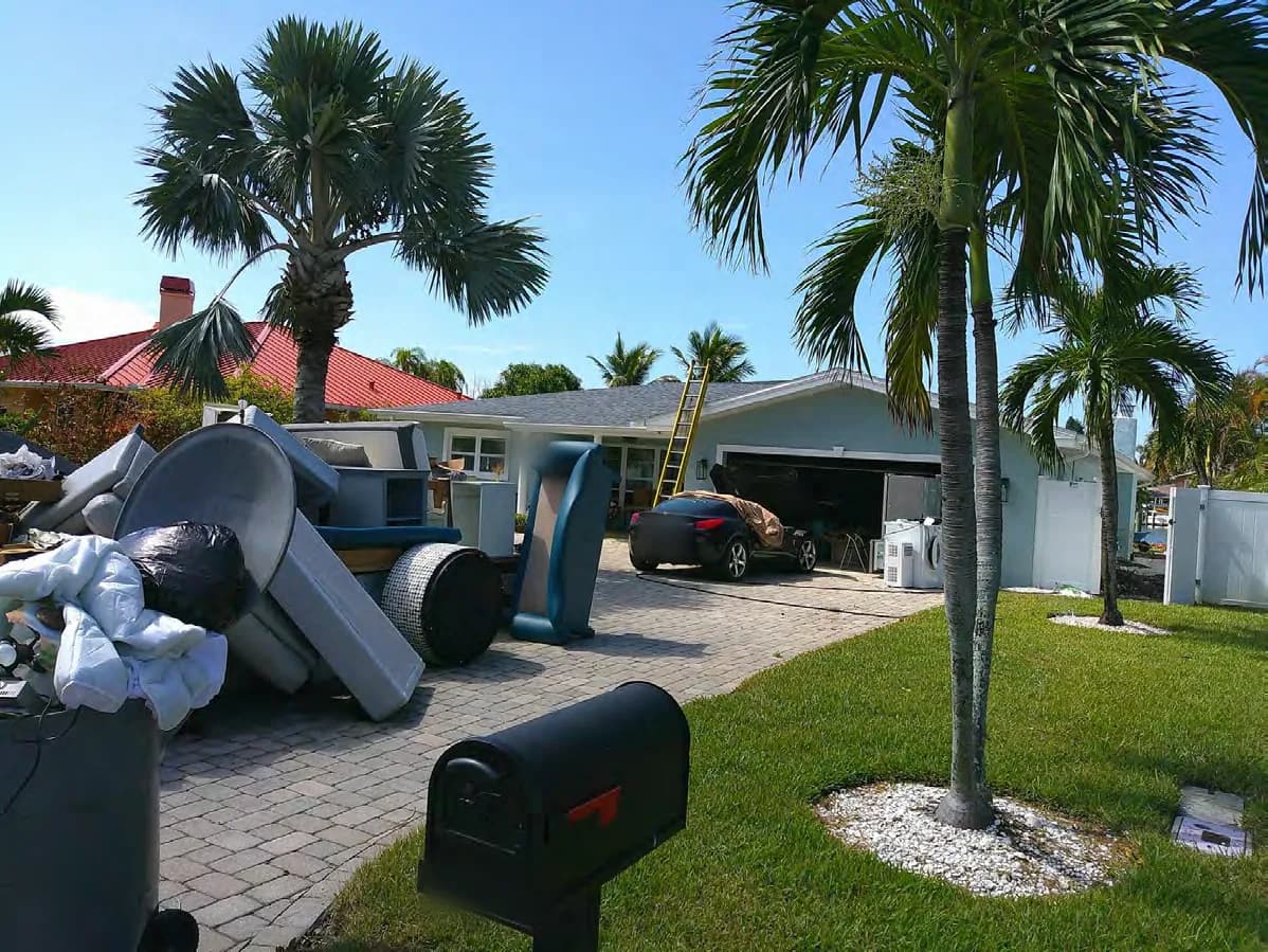 Water-damaged furniture and belongings piled at the curb of a single-story home, with a ladder set against the roof and the garage standing open.
