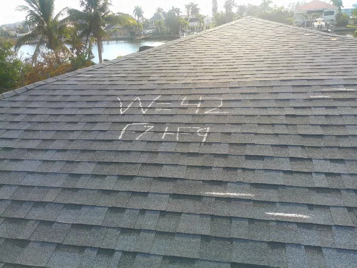 Grey shingle roof slope with a wind-damage tally written in chalk on the shingles and a canal behind the house.