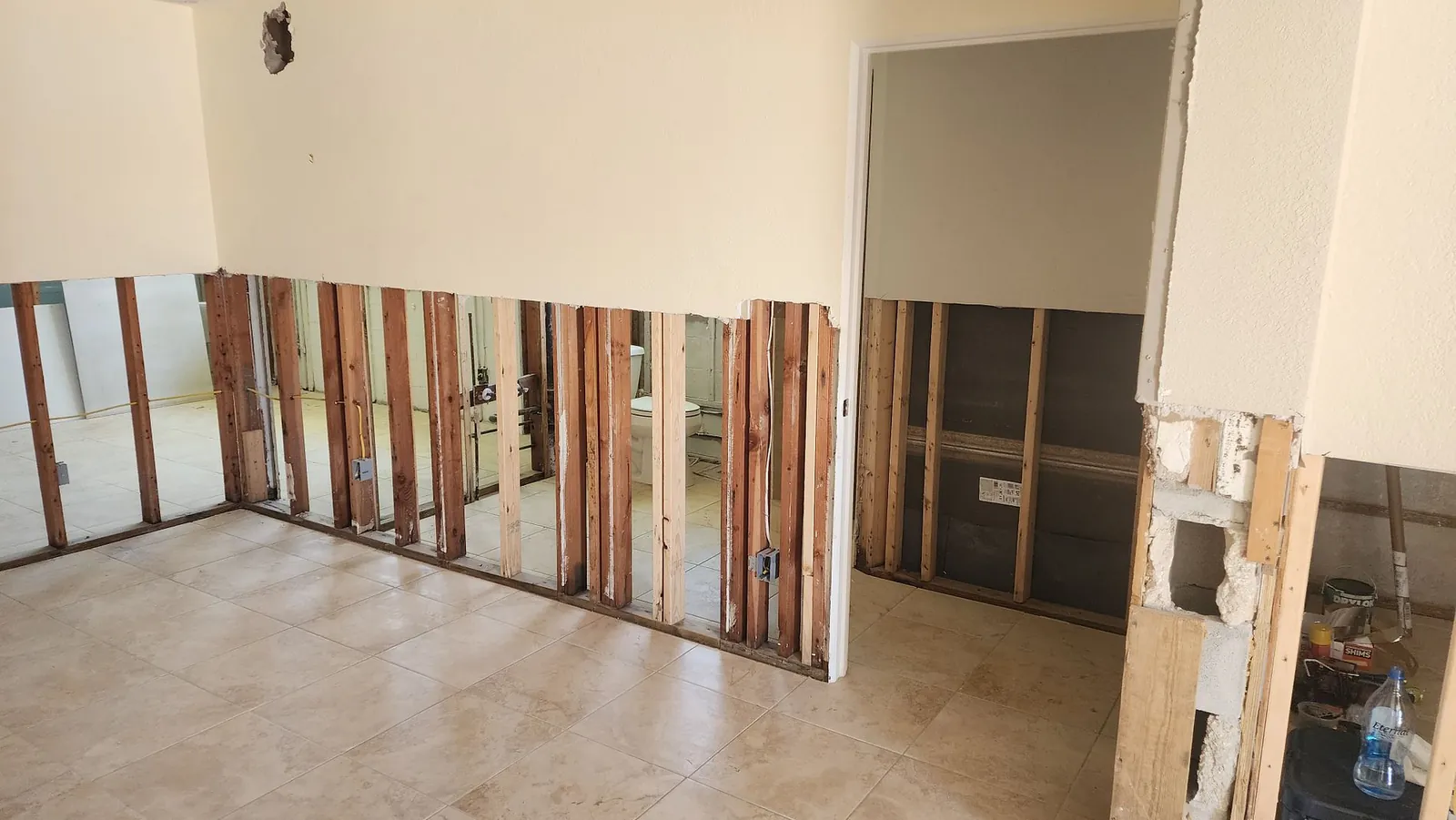 Interior room with the lower four feet of drywall removed to bare studs on every wall, tile floors exposed, and a bathroom visible through the open framing.