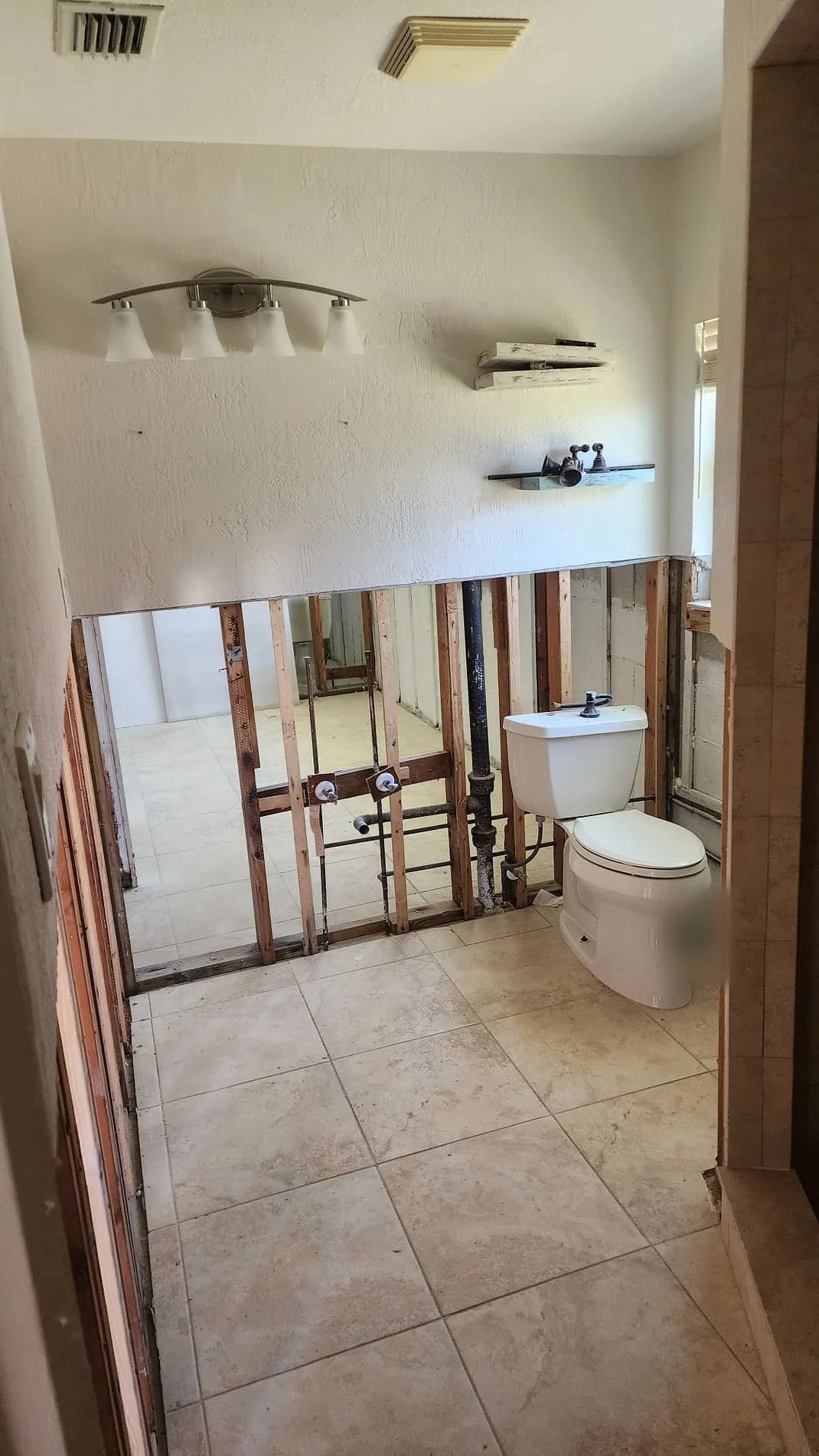 Bathroom with the lower wall cut away to the framing around the toilet, supply plumbing exposed, and the vanity light still mounted above.