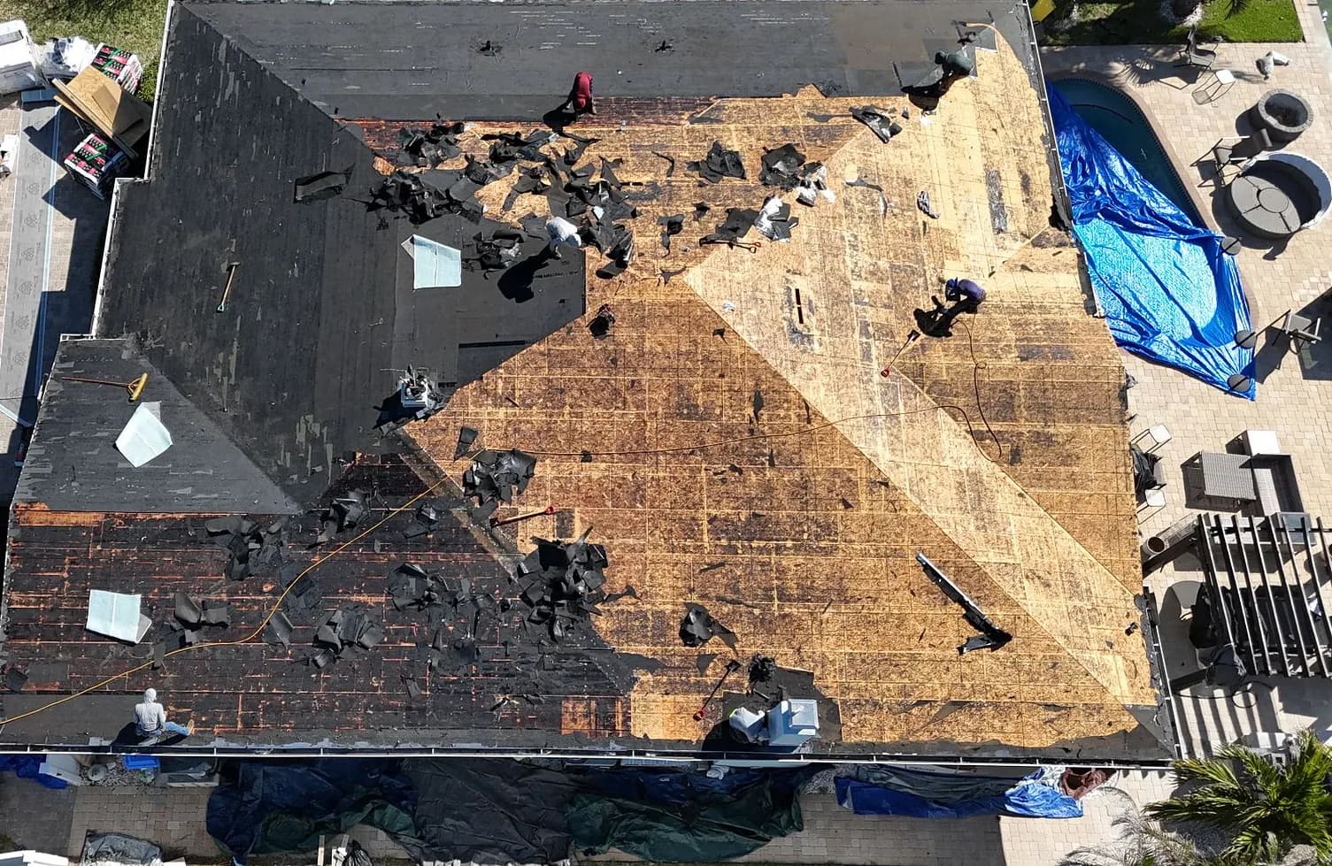 Aerial view of the roof mid-tear-off, one side stripped to bare sheathing, crew members working on the slopes, and tarps protecting the ground below.