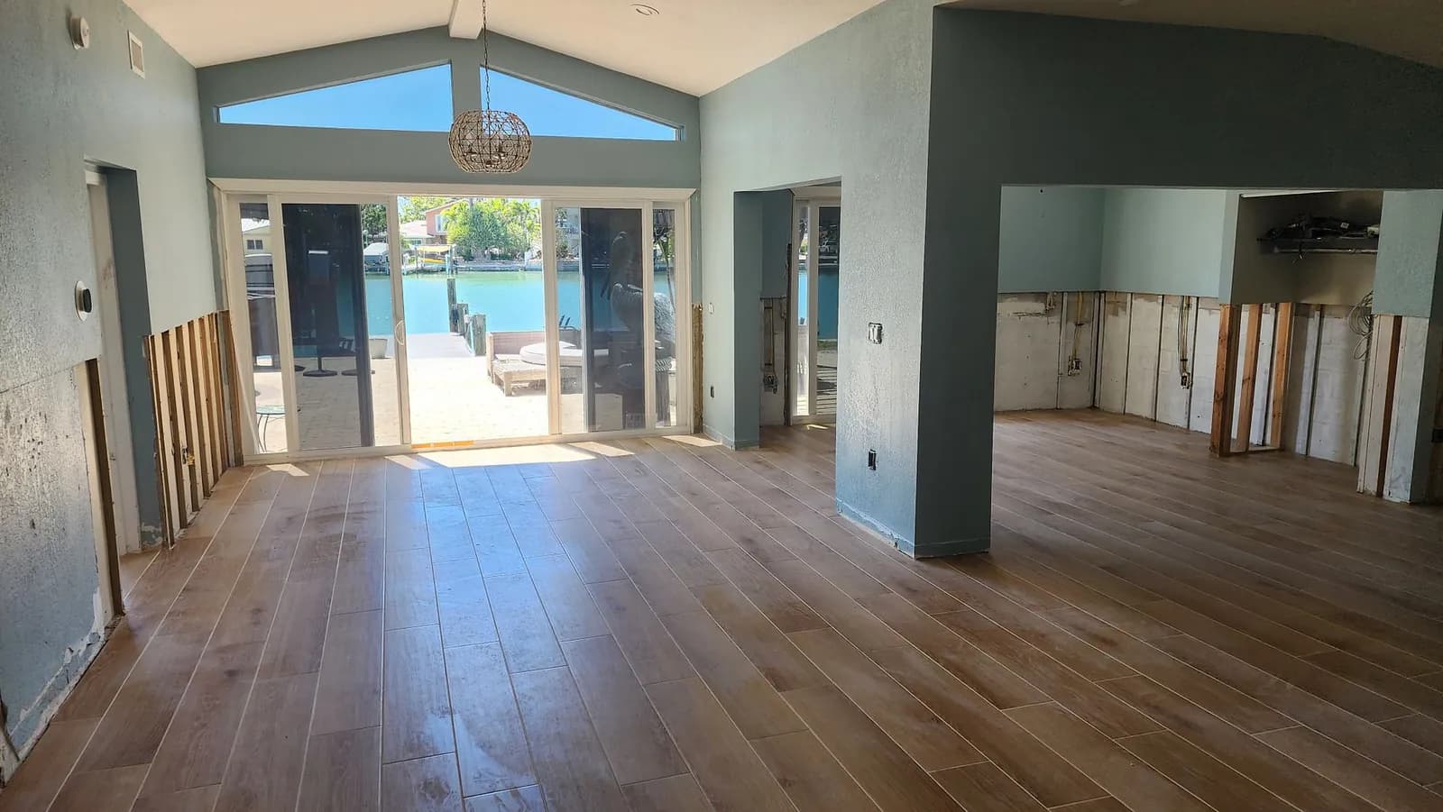 Open living space with new plank flooring installed, the base of the walls still open from the flood cut, and sliding glass doors looking out to a patio, dock, and canal.