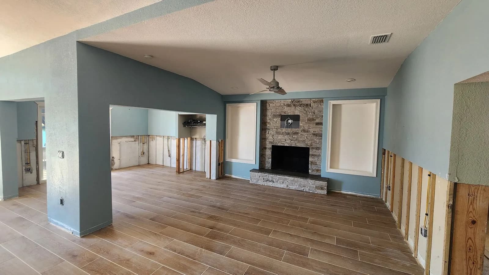 Living room mid-restoration with fresh paint and new plank flooring, a stacked-stone fireplace at center, and the lower walls still open to the framing.