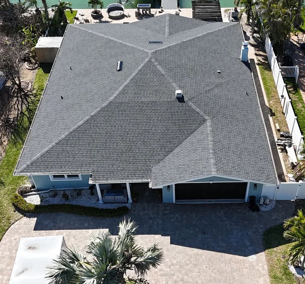 Overhead aerial of the completed hip roof in charcoal shingles, with the driveway in front and the canal and dock behind the house.
