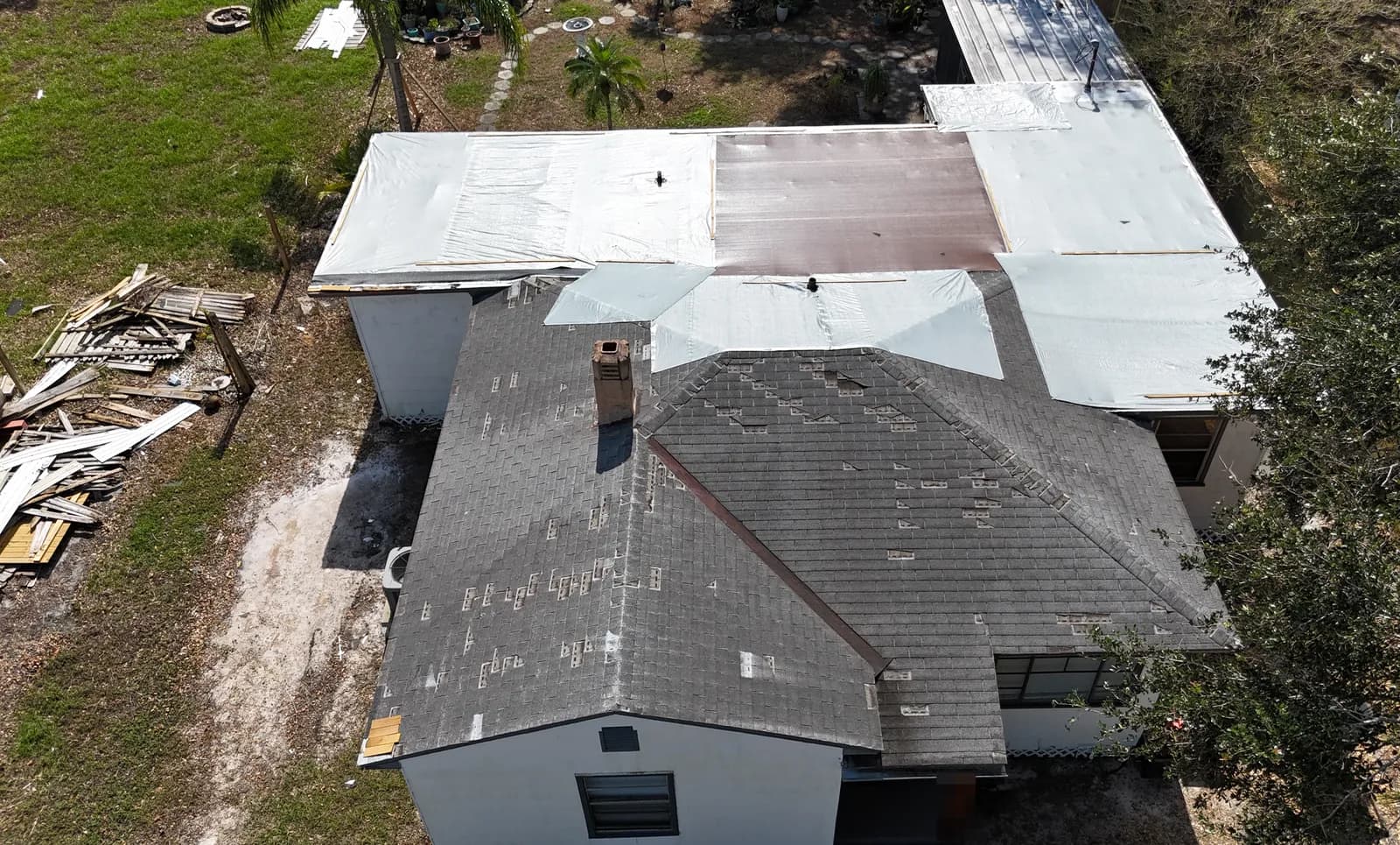Aerial view of a storm-damaged roof with shingles stripped on every slope, the flat section covered in tarps and membrane, and debris piled in the yard