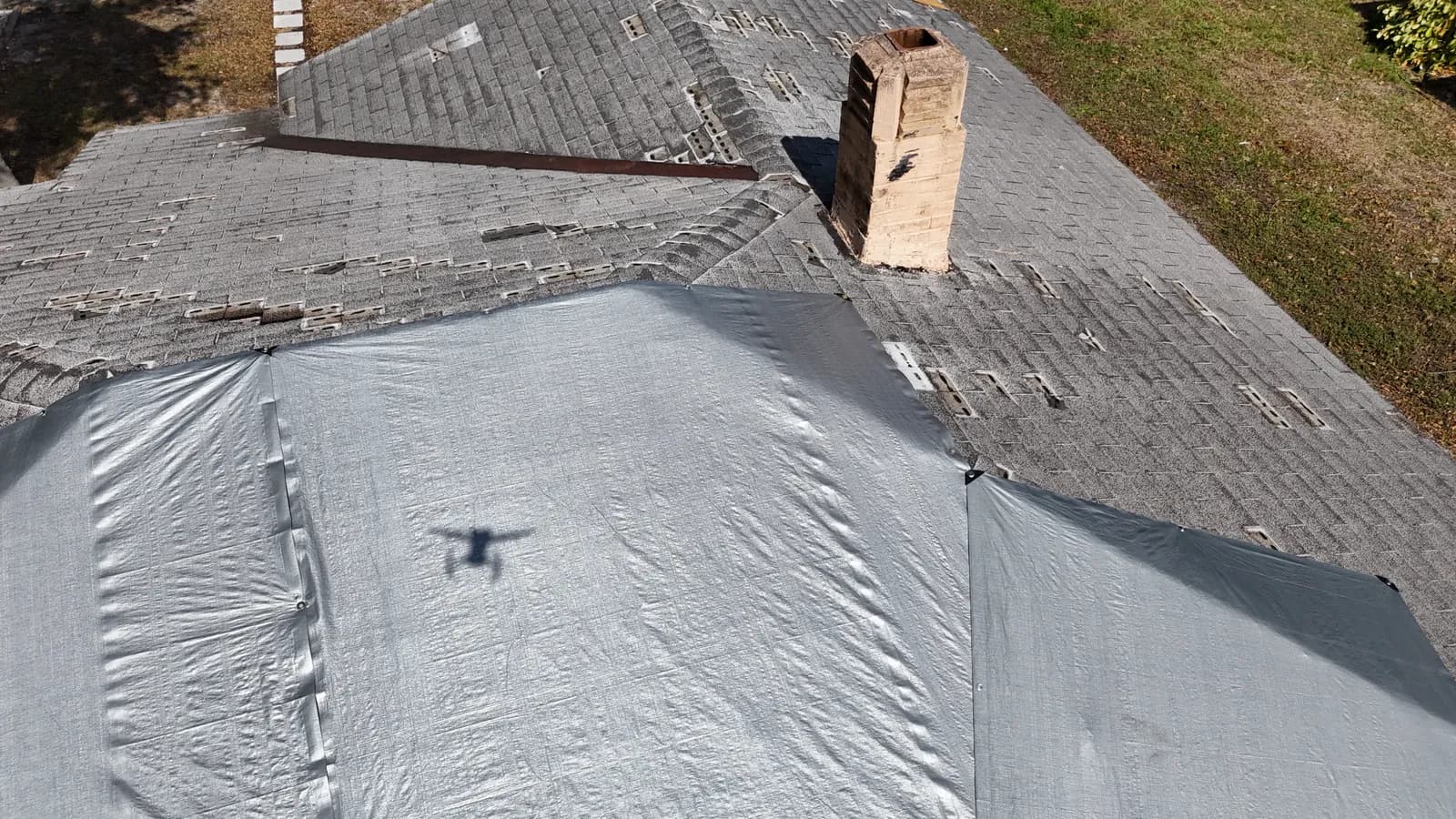 Close aerial view of shingle tab loss across a roof, a weathered brick chimney, and a gray tarp covering the lower slope