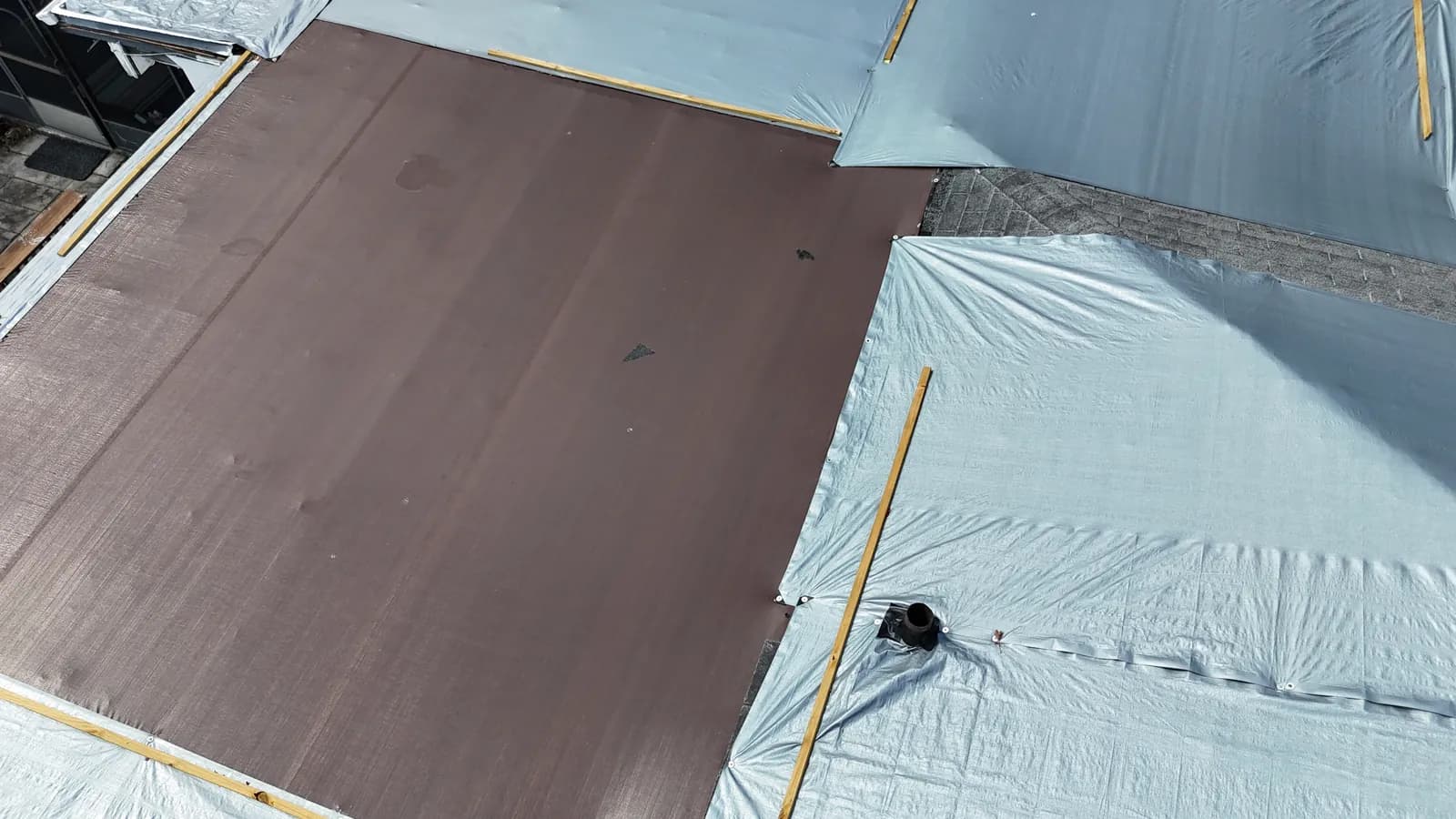 Overhead view of a flat roof section covered with brown peel-and-stick membrane, gray tarps, and wood battens