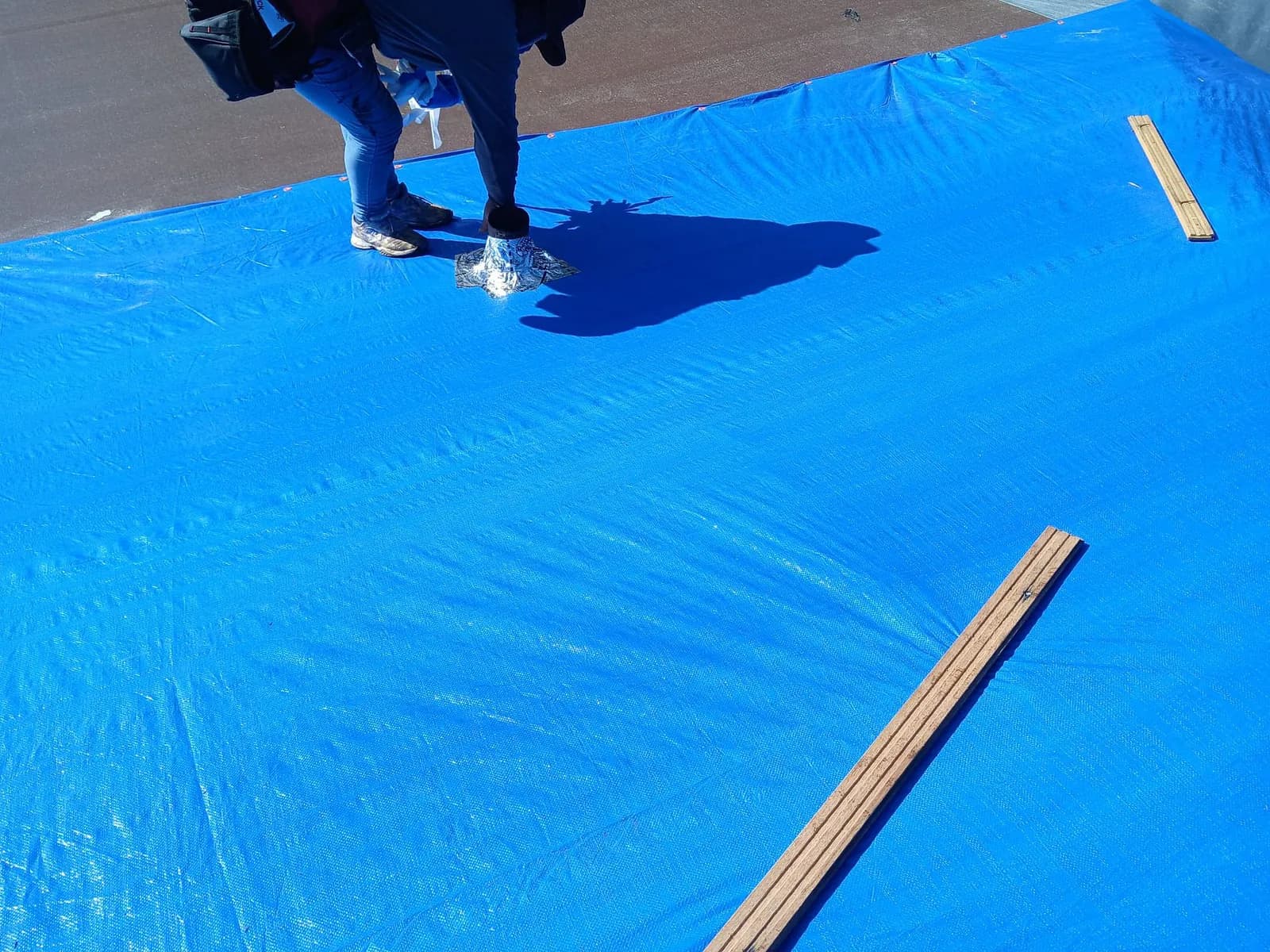Roofer standing on a fresh blue tarp stretched over a roof, with wood battens laid nearby