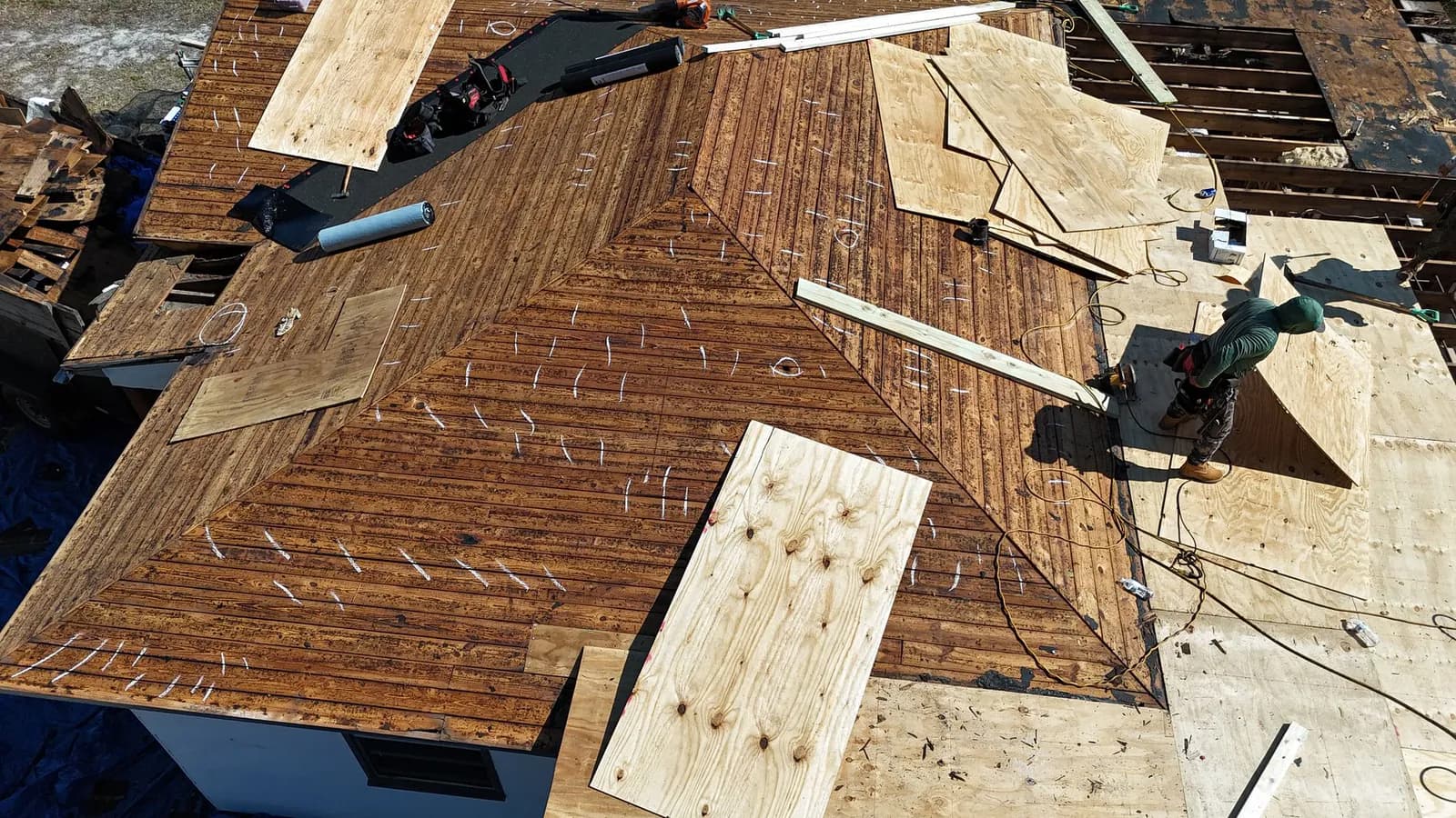 Aerial view of a roof mid rebuild, original plank decking exposed with new plywood sheets staged and a crew member working