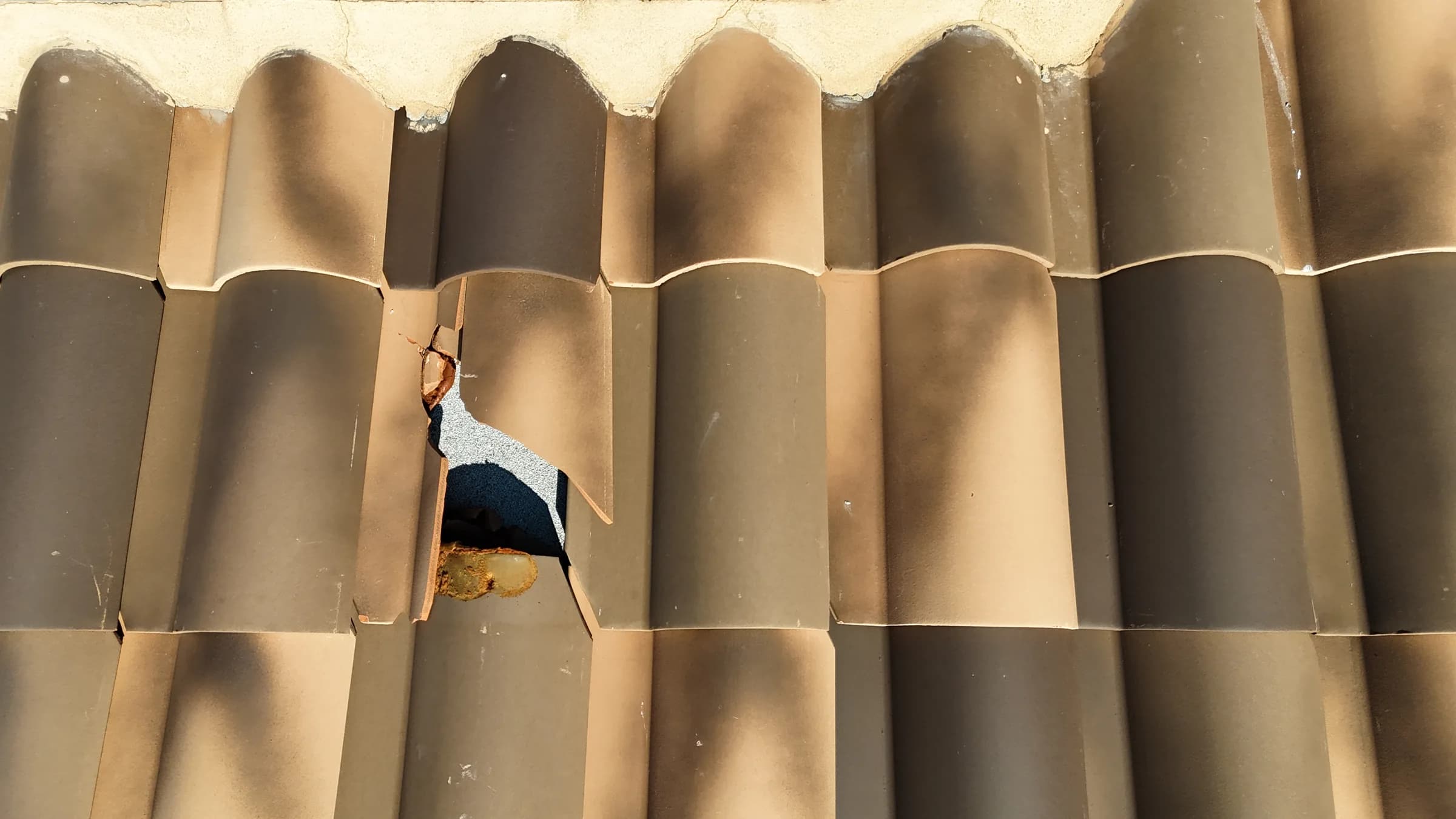 Close-up of broken brown barrel tiles with a hole exposing gray roof material below