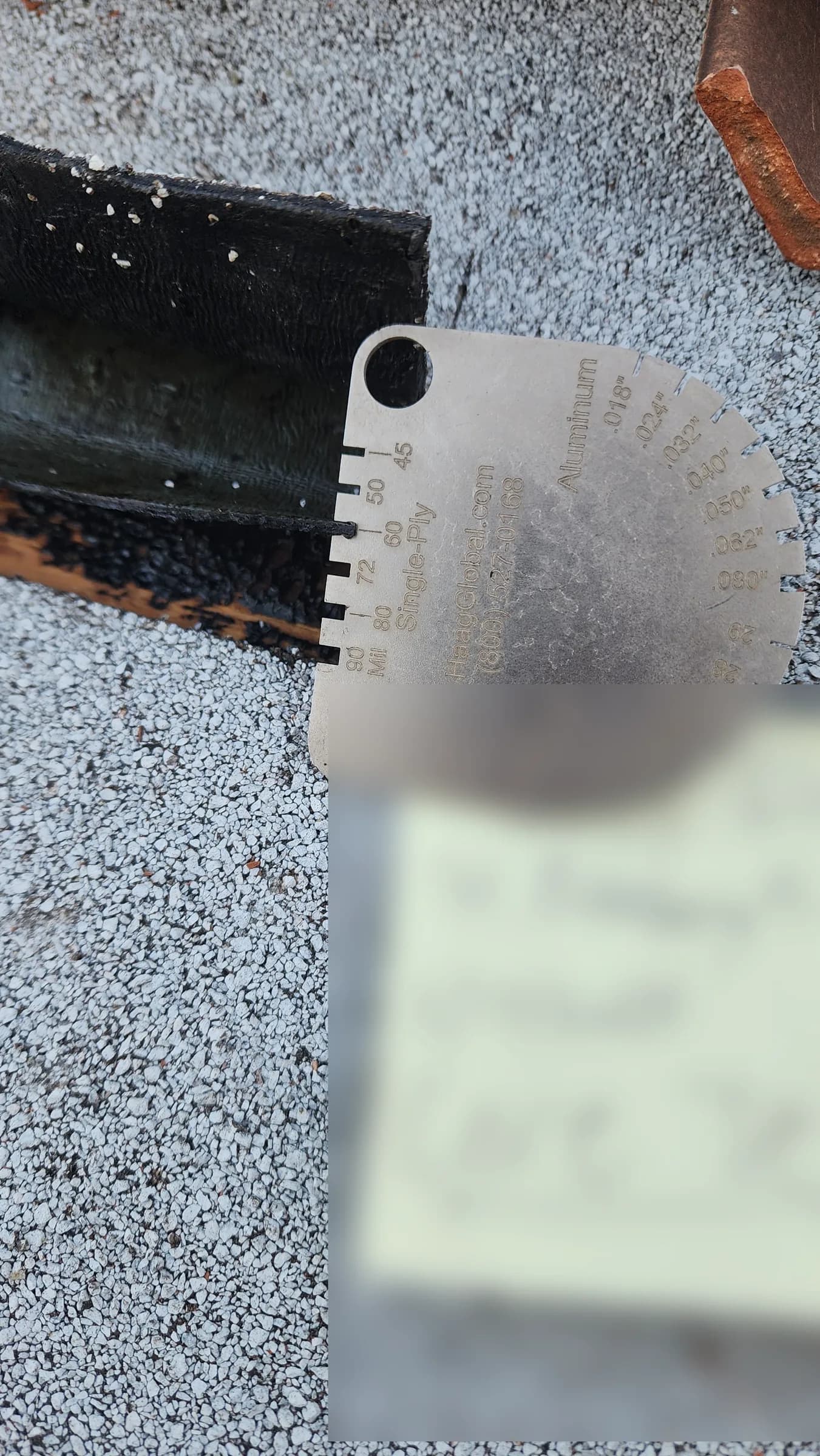 Thickness gauge placed against the black underside of a cut roof membrane sample on gray granulated underlayment