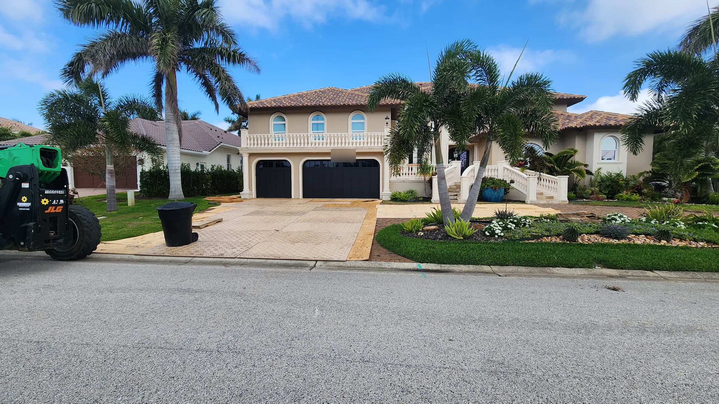 Two-story home with plywood and composite mats covering the clay brick driveway and a telehandler beside the curb