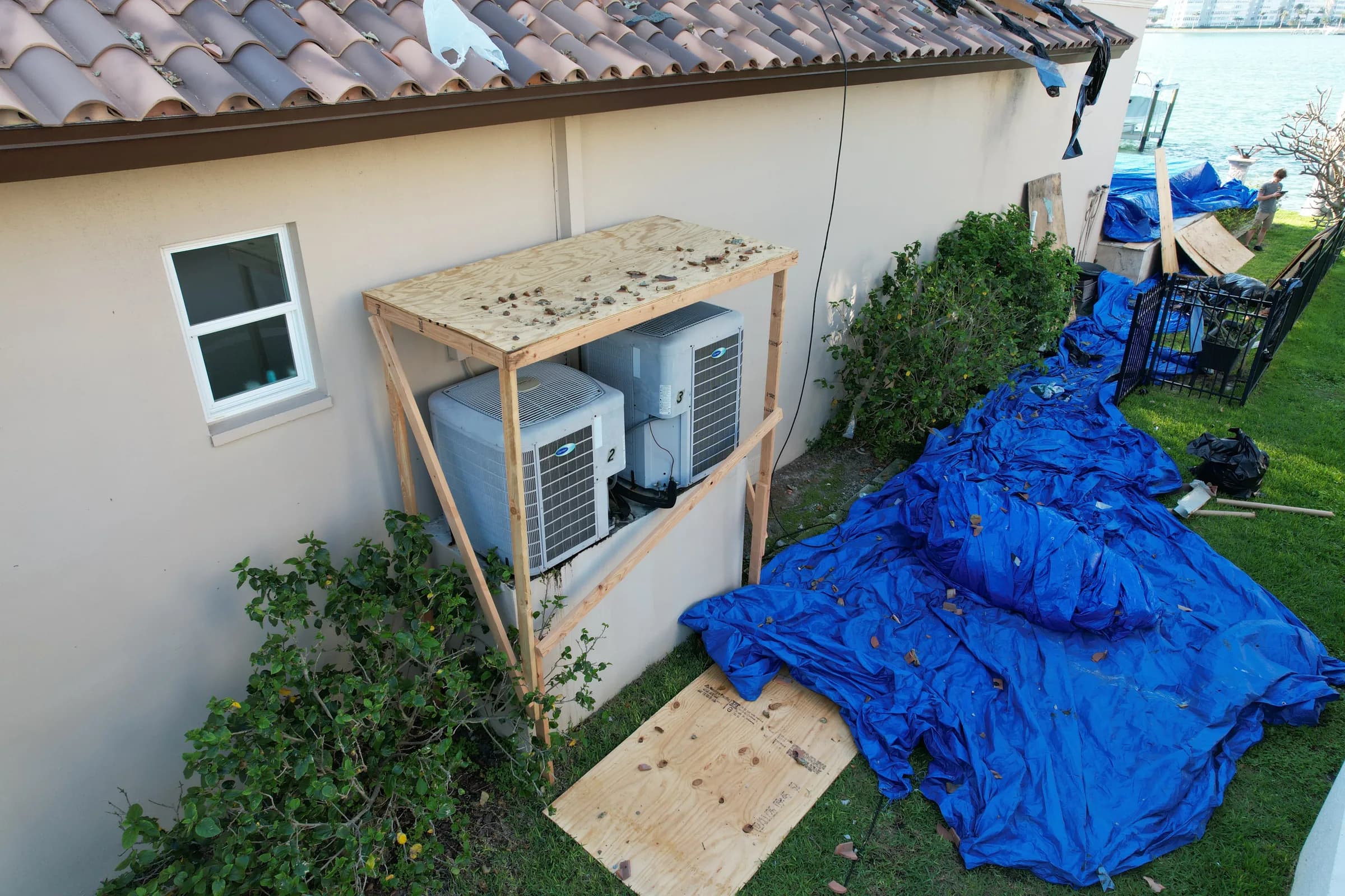 Plywood box built over two outdoor air-conditioning units with blue tarps covering the ground around the house