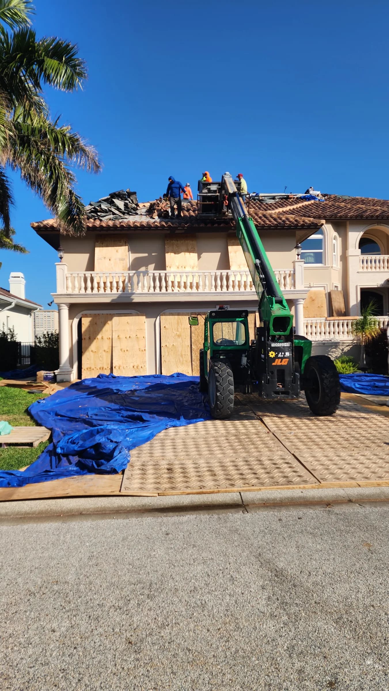 Roofing crew removing tile above boarded windows while a telehandler stands on the protected driveway