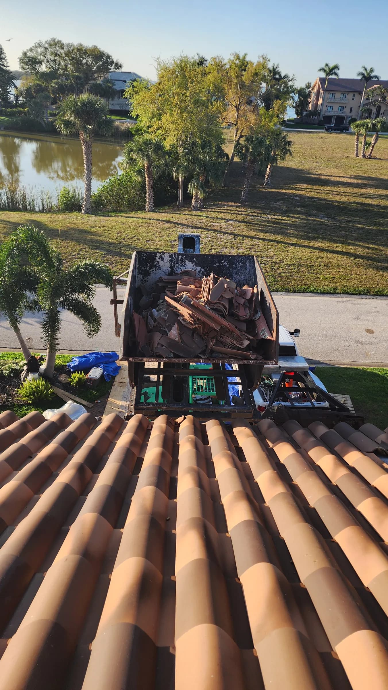 Roof-level view of a debris bin filled with removed clay tile beside the remaining tile field
