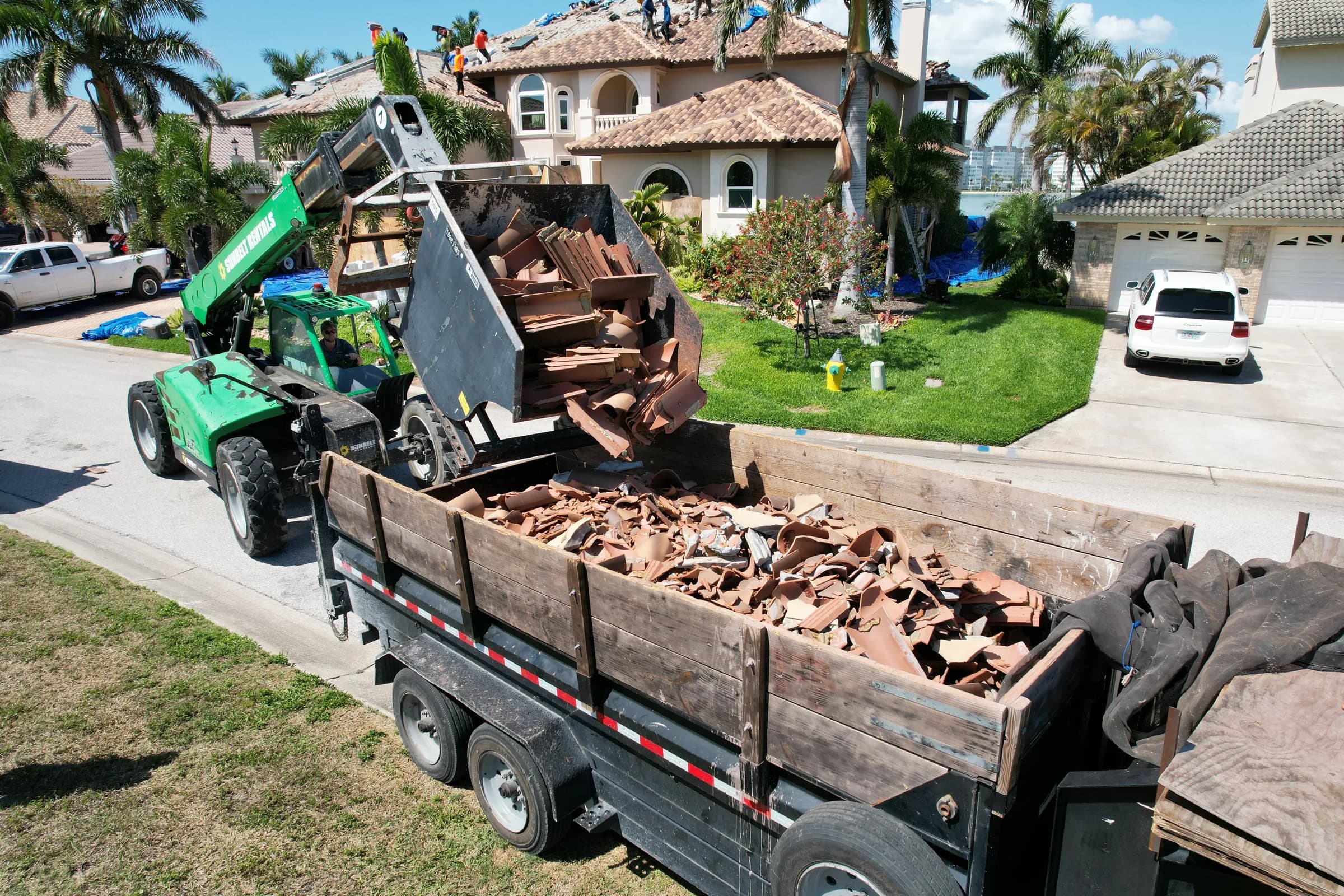 Telehandler tipping removed clay tile into a dump trailer while roofers continue tear-off on the house