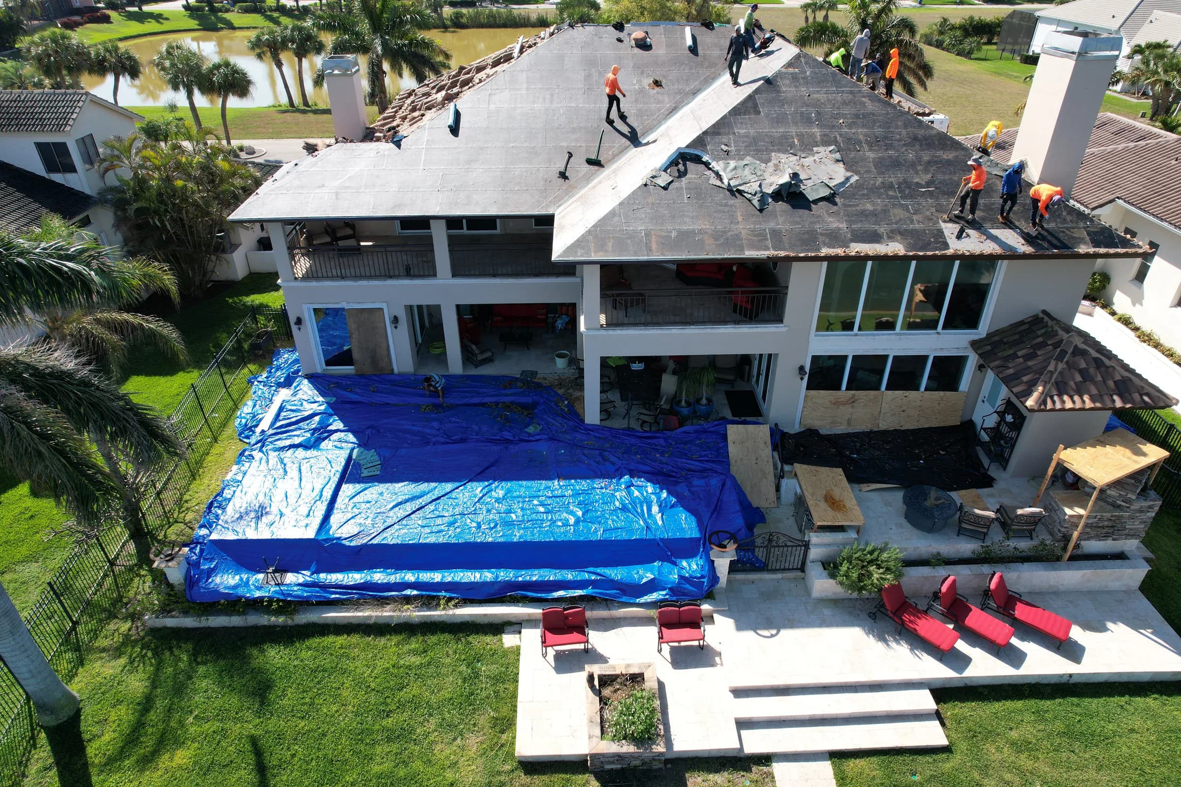 Aerial view of crews working across roof slopes stripped of tile with blue tarps covering the pool and patio below