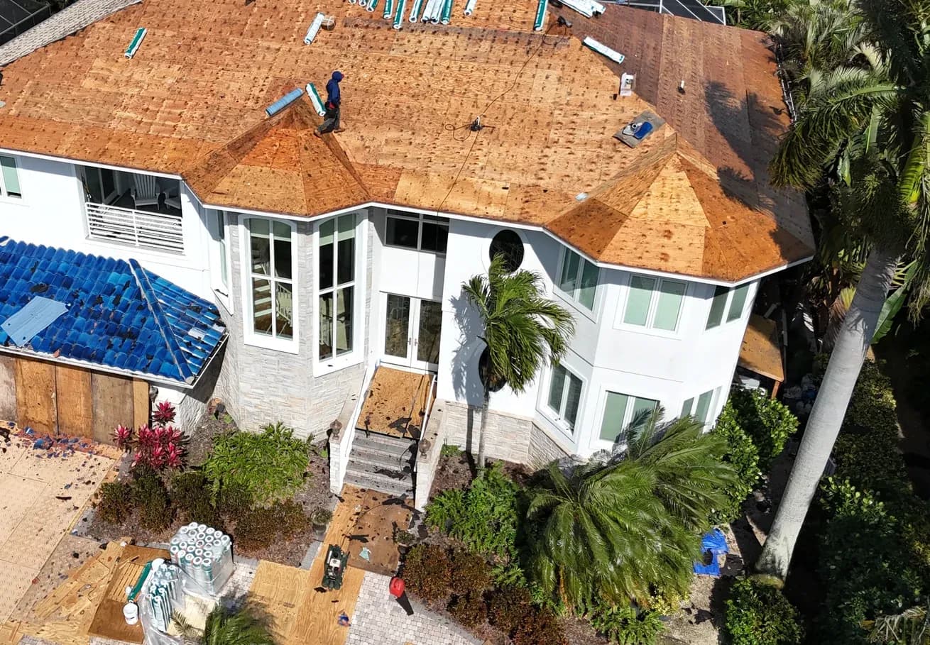 Aerial of the house with all tile removed and the roof deck bare, underlayment rolls staged on the plywood and on the driveway.