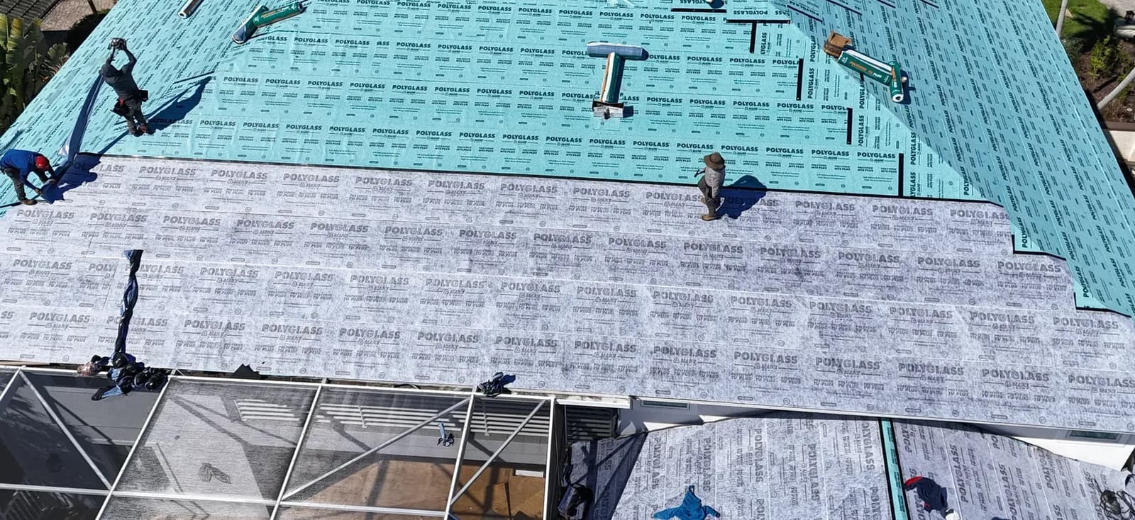 Roofing crew installing the two-ply underlayment across the roof, the gray cap sheet advancing over the teal base sheet.