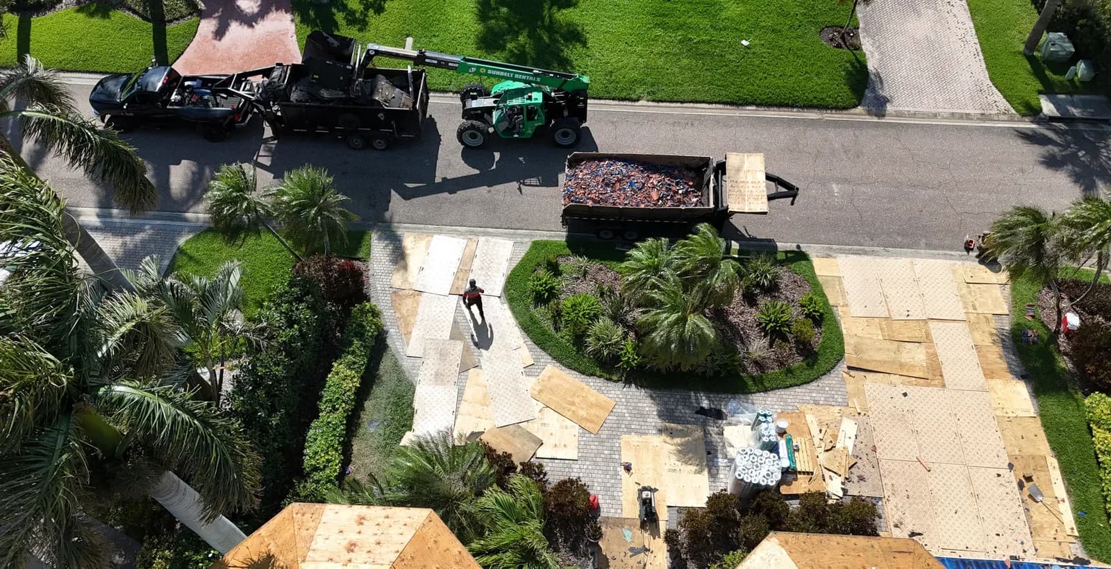 Overhead of a telehandler and a trailer loaded with broken tile at the street, with plywood ground protection across the paver driveway.
