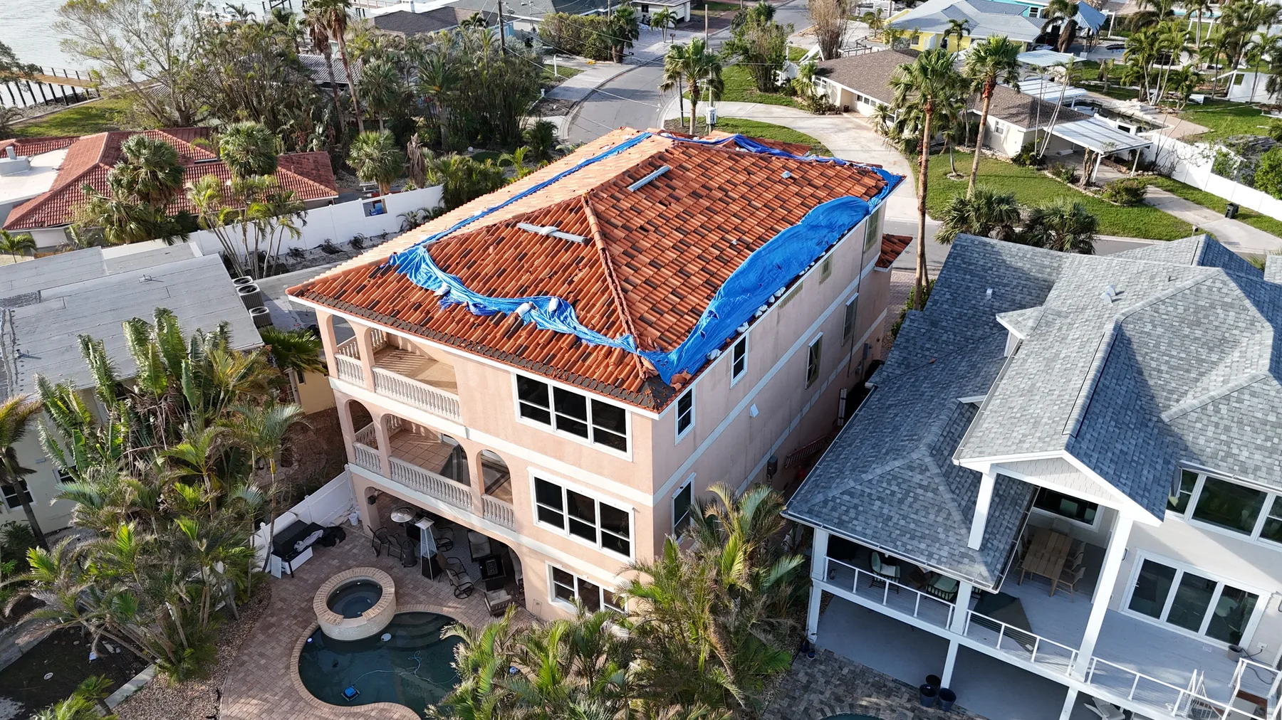 Aerial view of a three-story coastal home with a torn emergency tarp on its tile roof